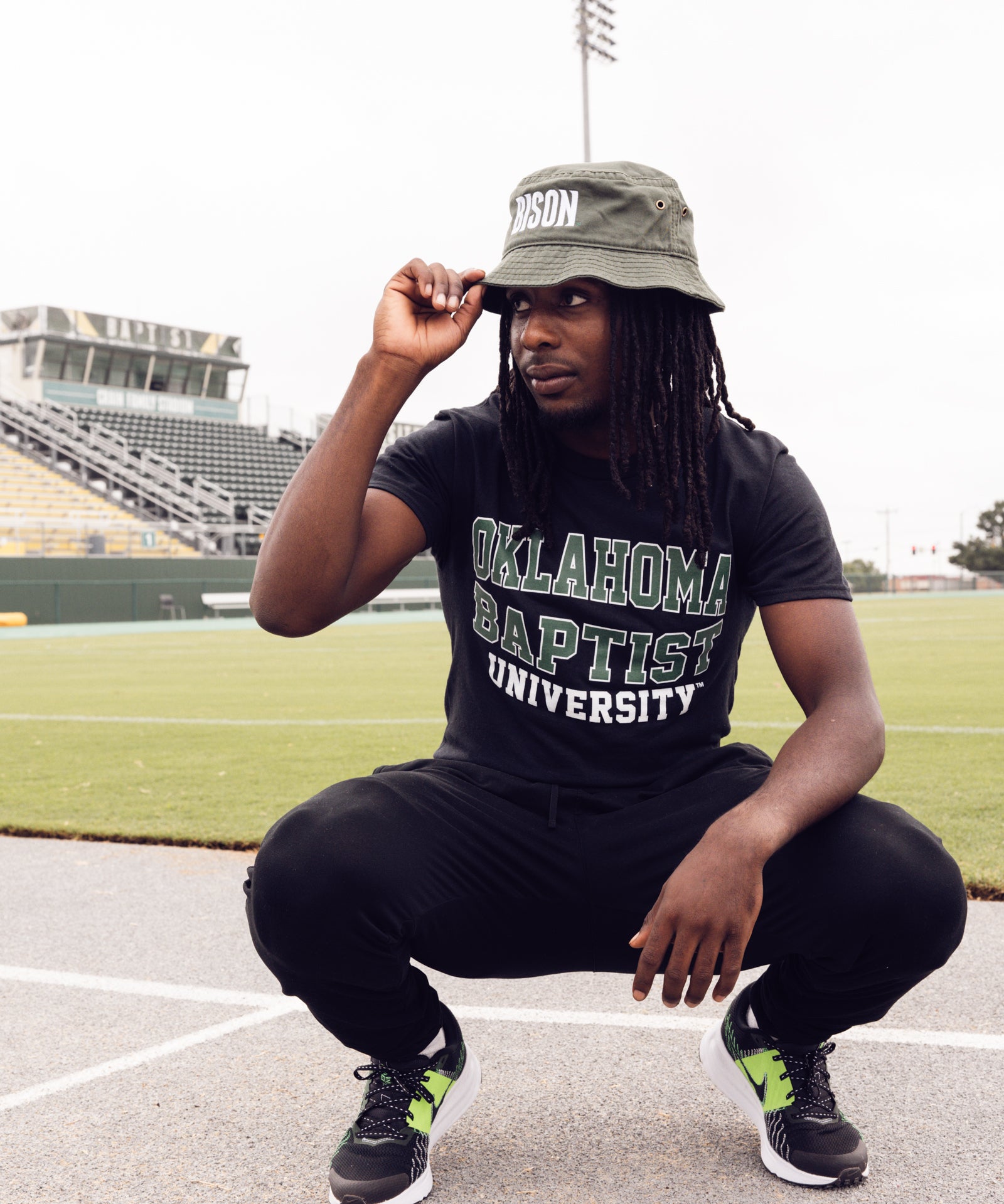 Person wearing an Oklahoma Baptist University shirt and a hat, squatting on a sports field.
