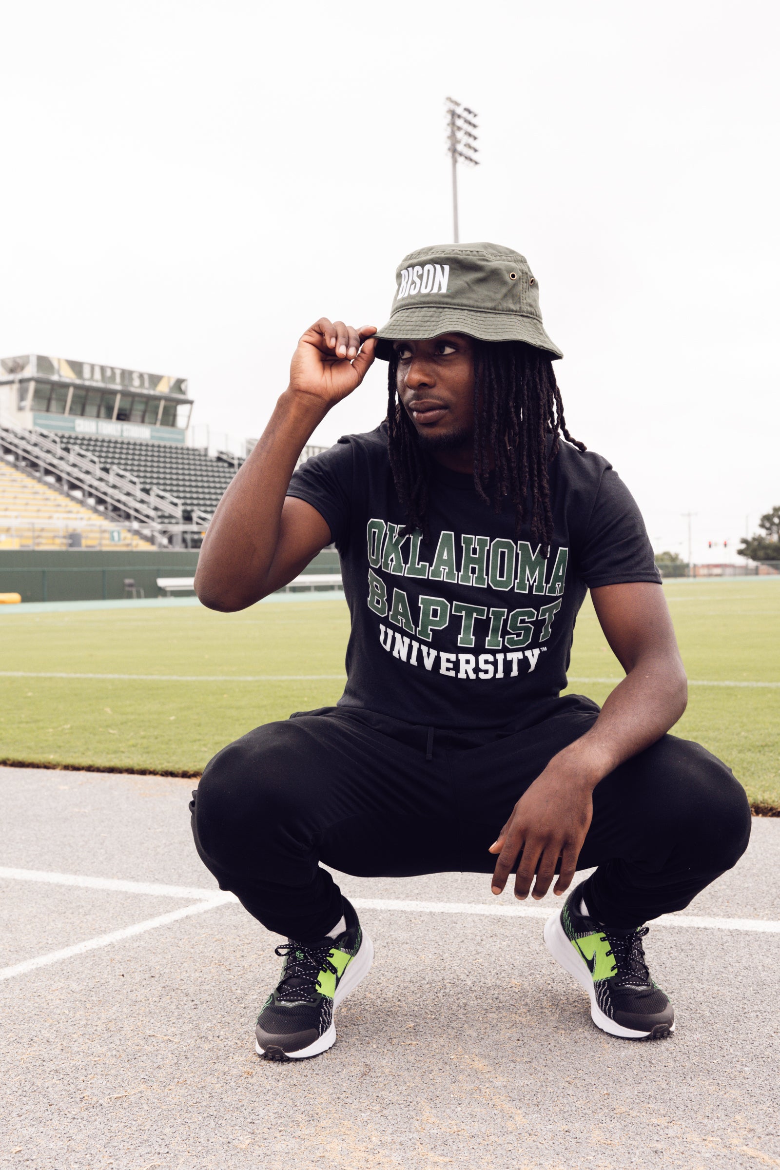 Person wearing an Oklahoma Baptist University shirt and a hat, squatting on a sports field.