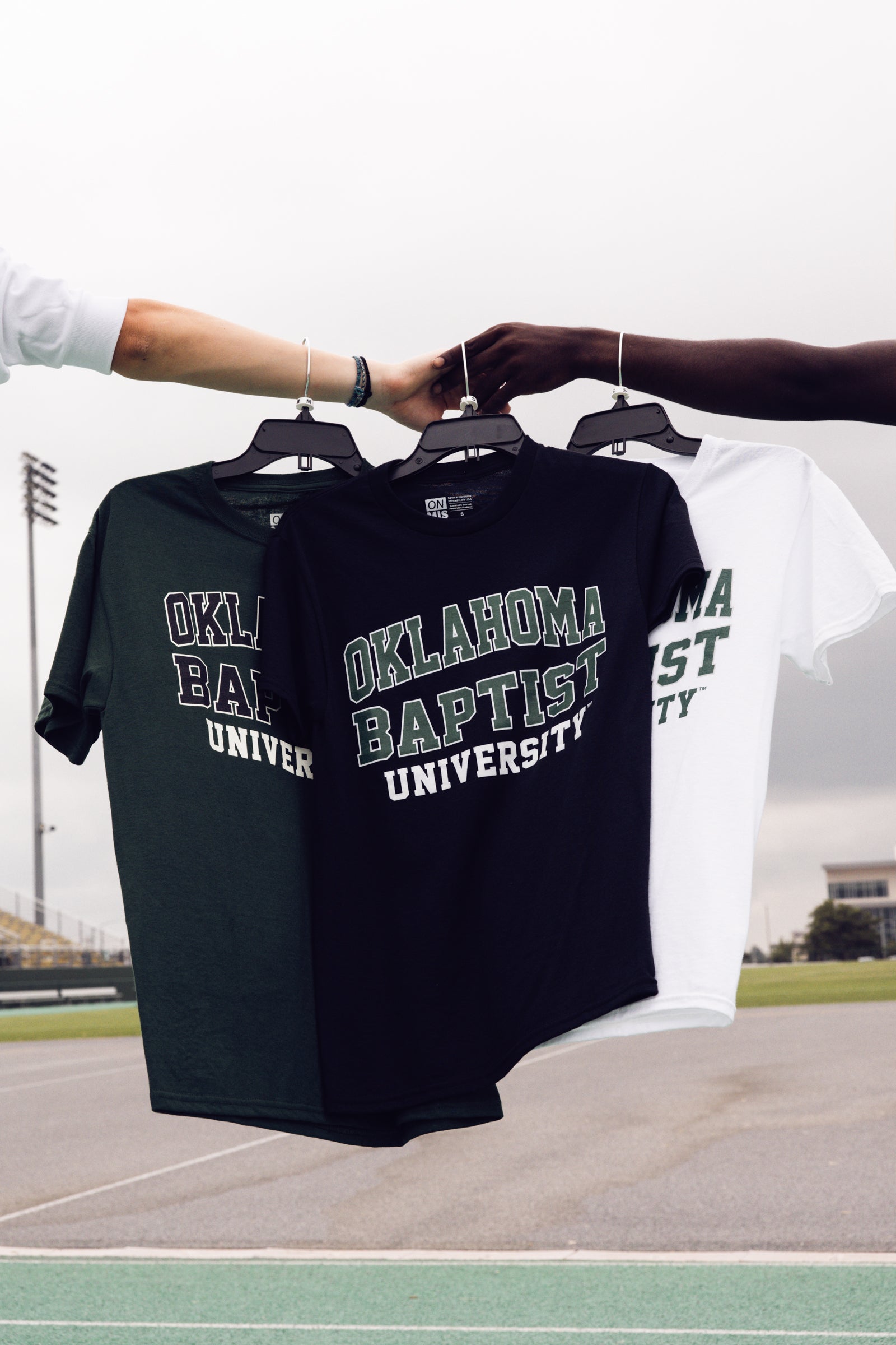 Two black t-shirts with 'Oklahoma Baptist University' printed on them, held by two people outdoors.