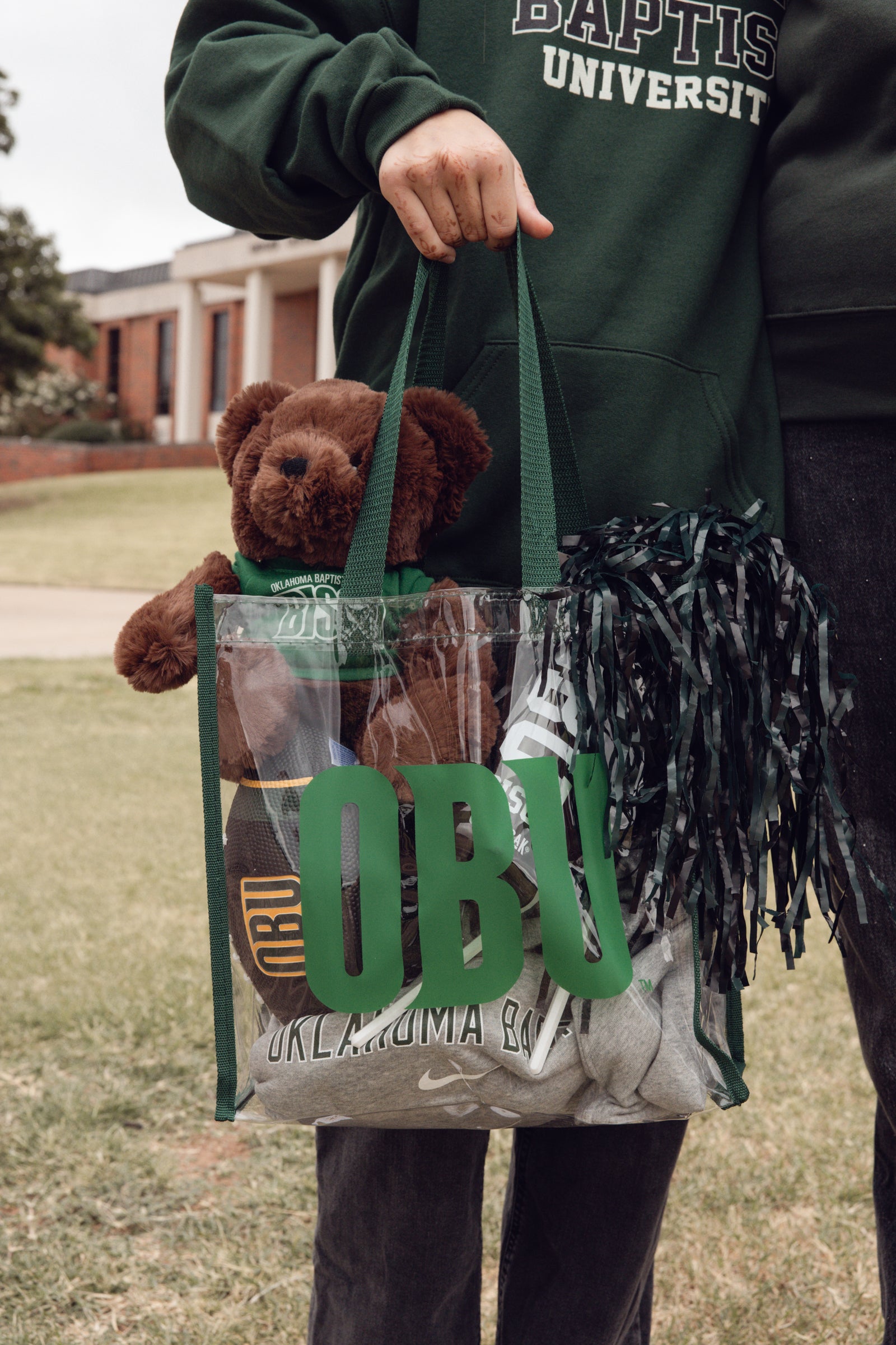 Person holding a clear bag with 'OBU' and a teddy bear, wearing a green sweatshirt with 'Baptist University' text.